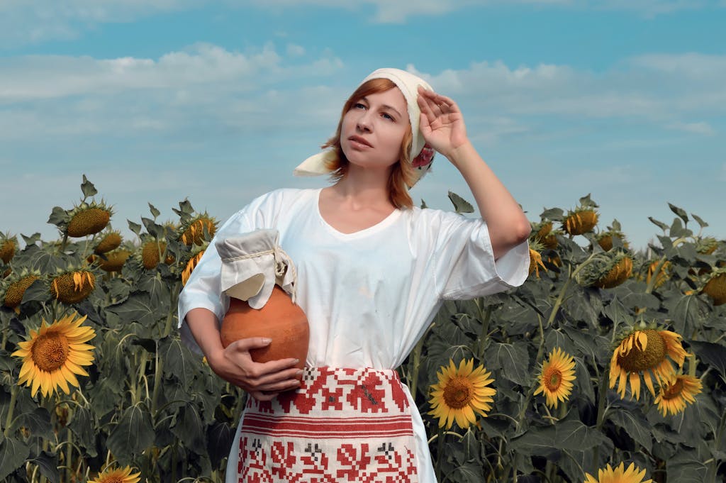 Young woman in traditional peasant country dress and embroidered apron holding earthenware jug in sunflower field in summer